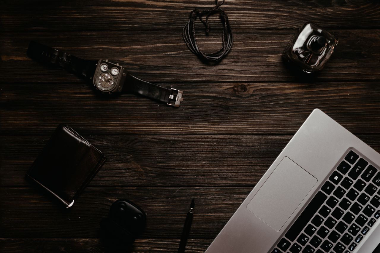 Flat lay of laptop, watch, wallet, and accessories on a dark wooden surface.